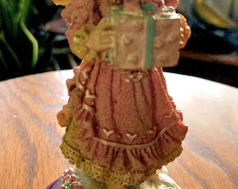 Może przedstawiać: Porcelanowa figurka anioła w różowej sukience z pudełkiem na prezent. Anioł ma blond włosy i niebieskie oczy. Figurka stoi na drewnianej podstawie.
