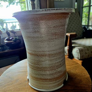 Könnte beinhalten: Ein hoher, strukturierter Keramik-Pflanztopf mit einer weiten Öffnung. Der Pflanztopf hat ein gesprenkeltes, cremefarbenes und braunes Design. Der Boden hat eine weiße, kreisförmige Stütze. Der Pflanztopf steht auf einem runden Holztisch.