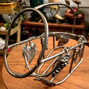 May include: Silver metal wine bottle holder with a grapevine design. The holder has a handle and is shaped like a basket.