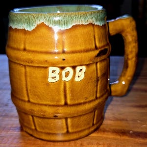 May include: A brown ceramic mug shaped like a barrel with a green glazed rim. The mug has the word "BOB" written on the front in white.