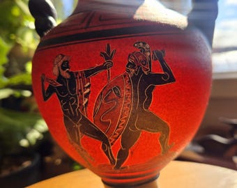Puede incluir: Un jarrón de cerámica rojo y negro con dos asas. El jarrón está decorado con una imagen en blanco y negro de dos guerreros griegos luchando. El jarrón tiene una base negra con un patrón geométrico rojo y negro.