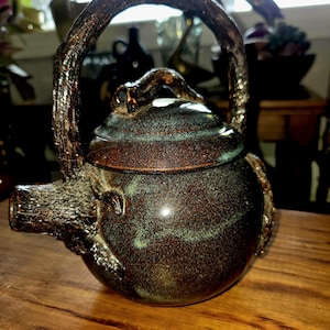 Puede incluir: Una tetera de cerámica marrón oscuro y verde con un mango y un pico únicos en forma de rama de árbol. La tetera tiene un aspecto texturizado y rústico.