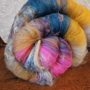 May include: A colorful, swirled bundle of fiber in shades of teal, yellow, pink, and tan. The fiber appears soft and fluffy, ideal for spinning or other textile crafts. The colors blend together in a gradient effect.