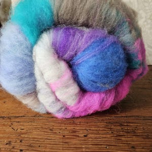May include: A close-up of a colorful ball of yarn. The yarn is a mix of blues, purples, pinks, and gray. The yarn is soft and fluffy, and it appears to be made of wool or a similar fiber. The ball of yarn is sitting on a wooden surface.