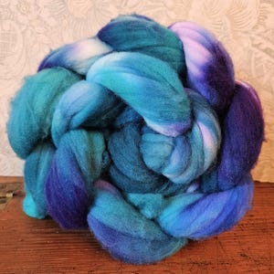 May include: A vibrant bundle of dyed wool roving in shades of teal, turquoise, and deep indigo. The fiber is arranged in a braided or coiled form, showcasing the color variations. Ideal for fiber art projects like spinning or felting.