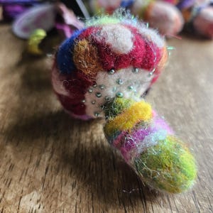 May include: A colorful, handmade felt mushroom ornament. The mushroom cap features a mix of red, white, blue, and orange hues, adorned with small, silver beads. The stem is a blend of yellow, green, and purple. The ornament is set against a wooden surface.