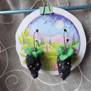 May include: A pair of handmade earrings shaped like strawberries. The earrings are black with small pink and iridescent accents, topped with green felt leaves. They hang from gold-colored ear wires and are displayed on a card with a whimsical illustration.