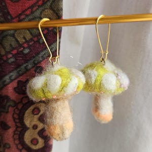 May include: A pair of whimsical mushroom earrings. Each earring features a yellow and white cap with a tan stem and orange accents. The earrings are suspended from gold-colored ear wires, perfect for adding a touch of nature-inspired charm to any outfit.