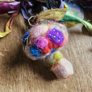 May include: A whimsical, handmade mushroom-shaped ornament. The cap features vibrant colors including pink, purple, red, and blue, with sparkling accents. The stem is a soft pink. The ornament is made of felted wool and other textiles.