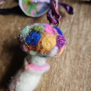 May include: A whimsical, handmade felt mushroom ornament. The mushroom cap is a vibrant mix of colors, including blue, pink, yellow, and green, with sparkly accents. The stem is white with a pink collar. A unique, decorative item.