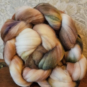 May include: A close-up of a braid of colorful wool roving. The roving is a mix of cream, brown, and green hues. The wool is soft and fluffy, perfect for fiber art projects like spinning or felting.