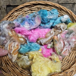 Könnte beinhalten: Ein Weidenkorb gefüllt mit bunten, flauschigen Fasern in Rosa-, Blau-, Gelb- und Brauntönen. Die Faser scheint handgefärbt zu sein und eignet sich zum Spinnen oder für andere Bastelprojekte. Der Korb besteht aus gewebtem, hellbraunem Material.