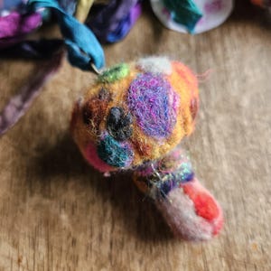May include: A handmade felted mushroom ornament with a colorful, textured design. The mushroom features a rounded cap with patches of purple, orange, green, and black. The stem is a mix of pink, red, and blue. Ribbons in various colors are in the background.