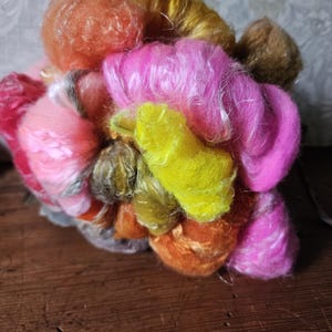 May include: A close-up of a bundle of colorful yarn in shades of pink, orange, yellow, and gold. The yarn appears soft and fluffy, with varying textures and thicknesses. The yarn is arranged in a compact, irregular shape.