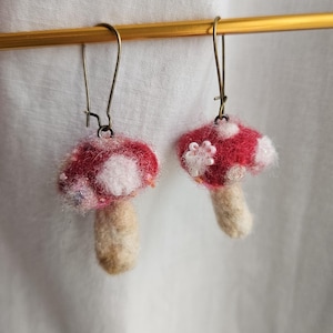 May include: A pair of handmade mushroom earrings. Each earring features a red cap with white spots and a beige stem. The earrings are suspended from bronze-colored ear wires, showcasing a whimsical, nature-inspired design.