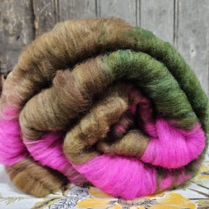 May include: A rolled bundle of colorful wool roving. The wool features shades of brown, green, and bright pink. The fibers are soft and fluffy, ideal for various fiber art projects like spinning, felting, and weaving.