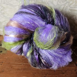 May include: A close-up of a vibrant ball of hand-dyed yarn. The yarn features a blend of purple, green, and brown hues, with strands of silver glitter. The texture appears soft and fluffy, ideal for knitting or other fiber crafts.