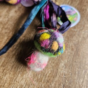 May include: Close-up of a handmade felt ornament with a colorful, speckled design. The ornament features a vibrant mix of yellow, pink, green, and blue, with a smaller white and pink felt ball attached. Dark blue and purple ribbons are attached.