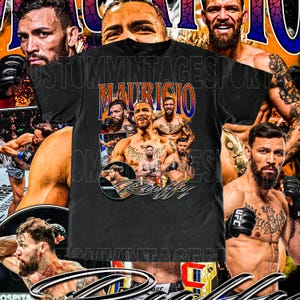 Peut inclure: T-shirt noir avec un collage d'images d'un combattant d'arts martiaux mixtes. Le t-shirt porte le nom "Mauricio" en lettres orange et bleu. Le mot "Ruffy" est en dessous des images.