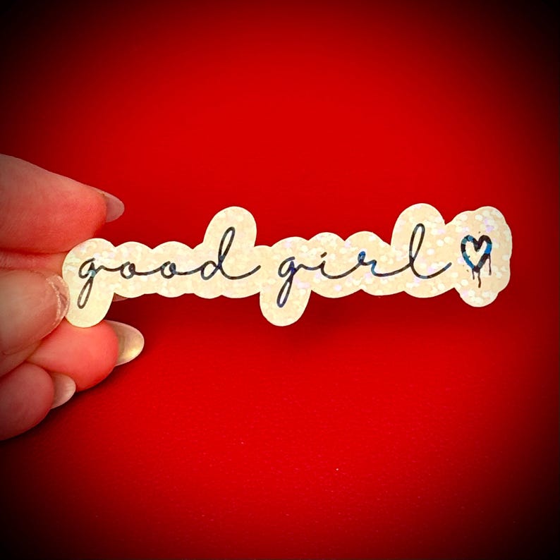 May include: A sticker with the words "good girl" in a handwritten font. The sticker has a glittery, off-white background and a small blue heart with a dripping effect. The sticker is held against a red background.