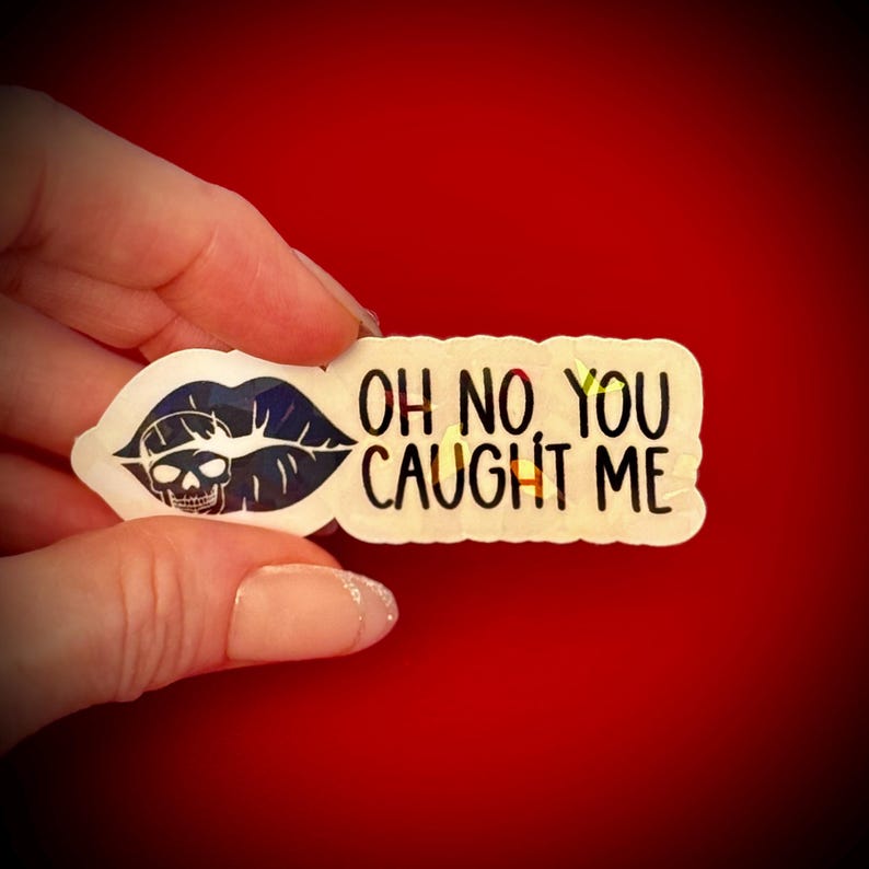 May include: A sticker with a skull inside a blue and black lip design, with the words "OH NO YOU CAUGHT ME" in black text. The sticker is held against a red background.