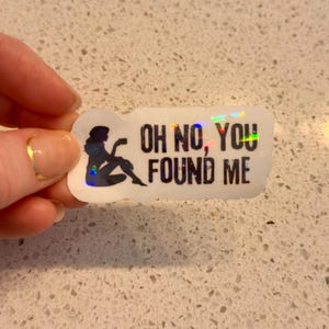 May include: A white sticker with the text "OH NO, YOU FOUND ME" in black. A silhouette of a person reading a book is on the left. The sticker has a holographic, iridescent finish.