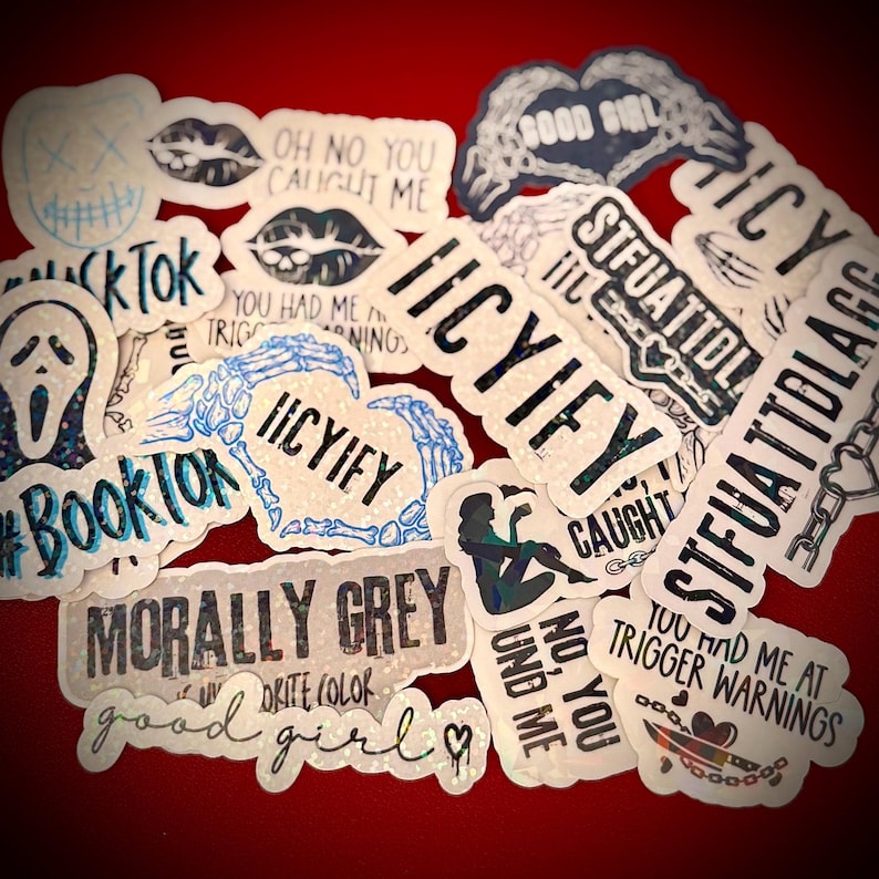 May include: A collection of holographic stickers with various designs and text. The stickers feature phrases like "ICYIFY," "MORALLY GREY," and "#BookTok." Other designs include lips, a ghost face, and a skeleton hand heart. The stickers are on a red background.