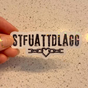 May include: A white sticker with the text "STFUATTDLAGG" in black, outlined in white. Below the text is a chain design with a heart in the center. The sticker has a holographic, glittery effect.