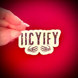 May include: A white sticker with the word "ICYIFY" in black letters with glitter accents and two skeleton hands. The sticker is held against a red background.