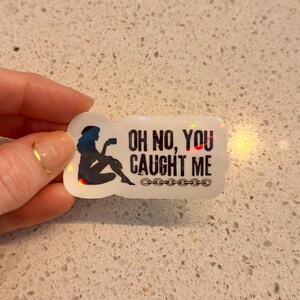 May include: A holographic sticker with the text "OH NO, YOU CAUGHT ME" and a silhouette of a person sitting and holding a cup. The sticker has a chain design at the bottom and is held by a hand.