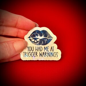 May include: A sticker with a skull inside a blue lip design. The sticker reads "YOU HAD ME AT TRIGGER WARNINGS" in black text. The sticker has a holographic finish and is held against a red background.