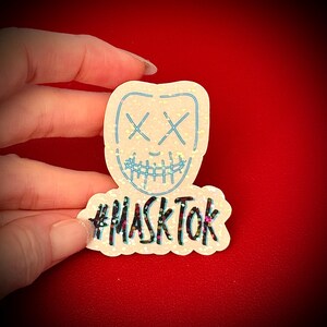 May include: A glittery sticker featuring a stylized mask design with crossed-out eyes and a stitched-up mouth, outlined in light blue. The text "#MASKTOK" is below the mask. The sticker is held against a red background.