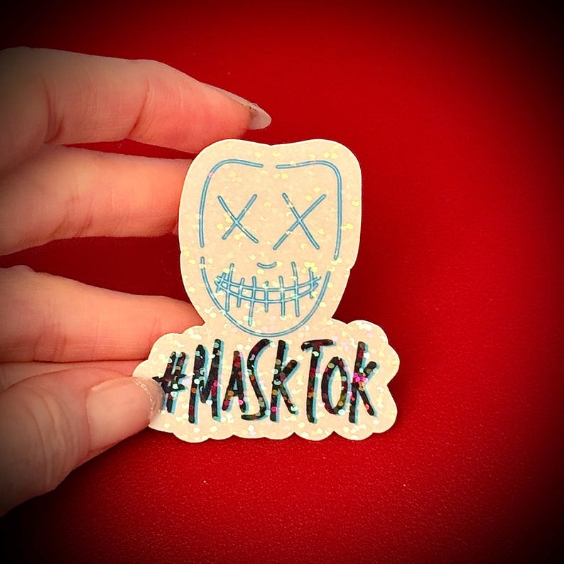 May include: A glittery sticker featuring a stylized mask design with crossed-out eyes and a stitched-up mouth, outlined in light blue. The text "#MASKTOK" is below the mask. The sticker is held against a red background.