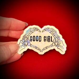 May include: A holographic sticker in the shape of a heart, featuring skeletal hands forming the heart shape. The words "GOOD GIRL" are printed in black within the heart. The sticker has a pearlescent sheen and is set against a red background.