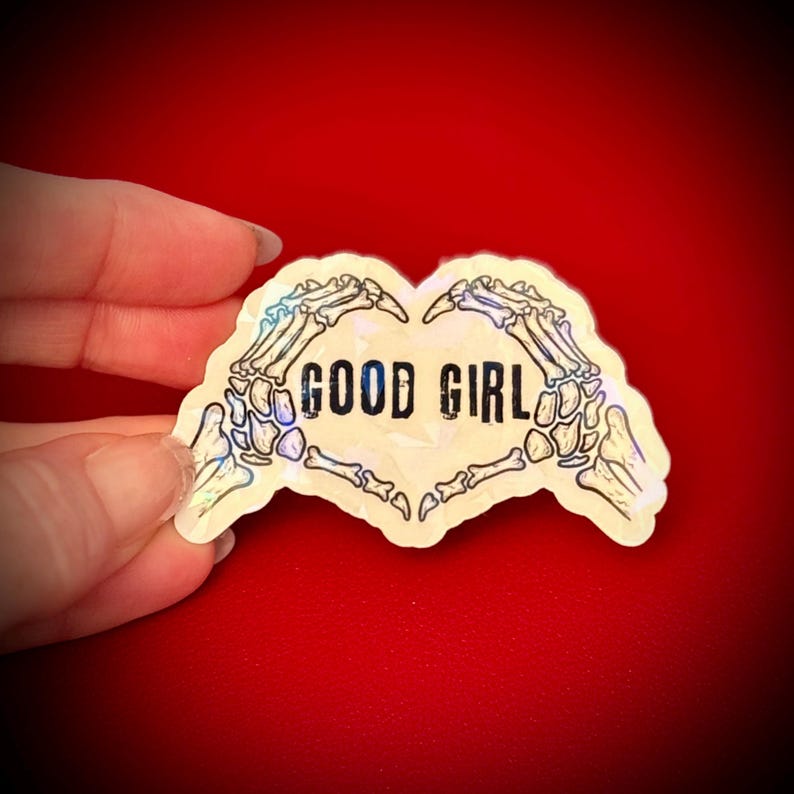 May include: A holographic sticker in the shape of a heart, featuring skeletal hands forming the heart shape. The words "GOOD GIRL" are printed in black within the heart. The sticker has a pearlescent sheen and is set against a red background.