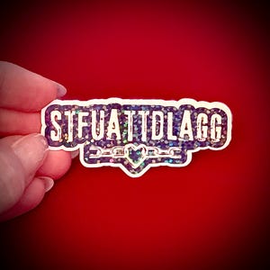 May include: A rectangular sticker with the text "STFUATTDLAGG" in white, outlined in a dark purple border. The sticker has a holographic, glittery effect. A heart and chain design is at the bottom. The sticker is held against a red background.