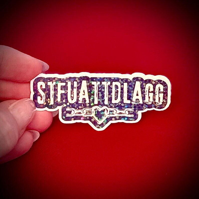 May include: A rectangular sticker with the text "STFUATTDLAGG" in white, outlined in a dark purple border. The sticker has a holographic, glittery effect. A heart and chain design is at the bottom. The sticker is held against a red background.
