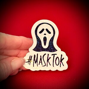 May include: A white sticker featuring the iconic Scream mask with a black interior and a glittery, dark blue outline. The text "#MASKTOK" is written in black below the mask. The sticker is held against a red background.