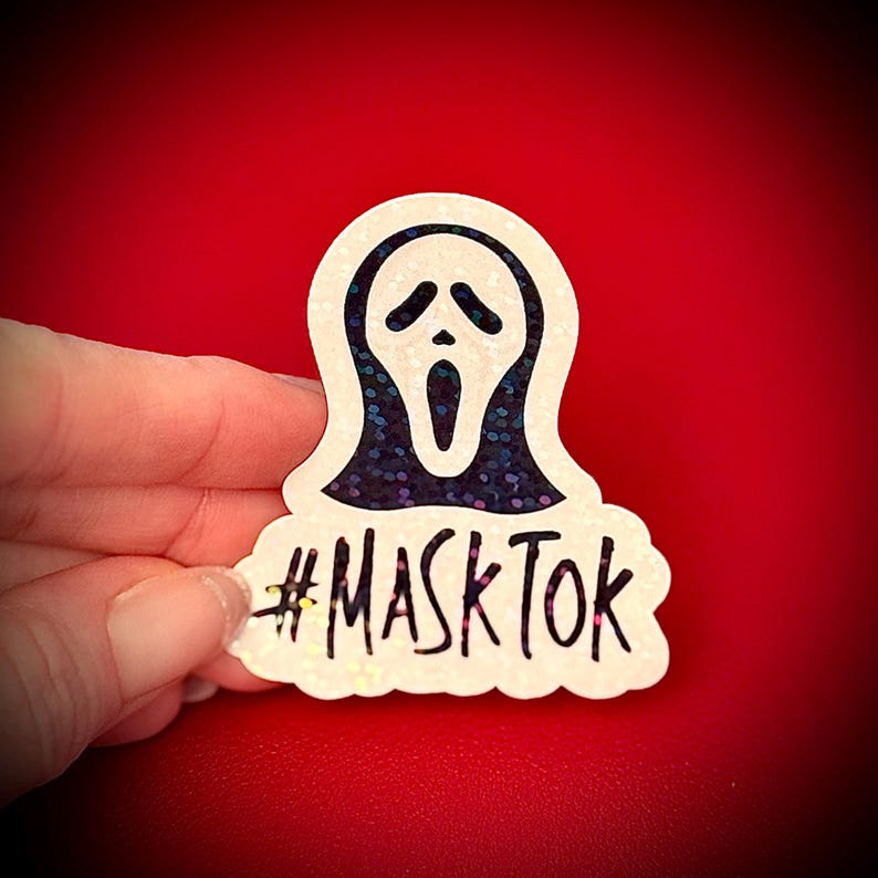 May include: A white sticker featuring the iconic Scream mask with a black interior and a glittery, dark blue outline. The text "#MASKTOK" is written in black below the mask. The sticker is held against a red background.