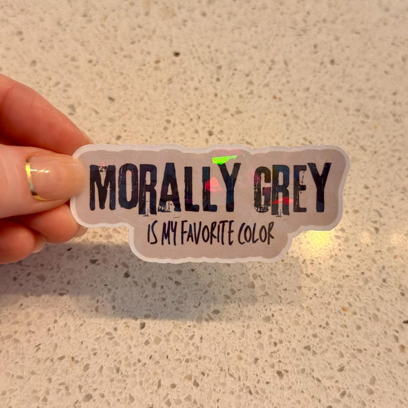 May include: A sticker with the text "MORALLY GREY IS MY FAVORITE COLOR" in a distressed font. The sticker has a light grey background with a holographic effect. The sticker is held by a hand.