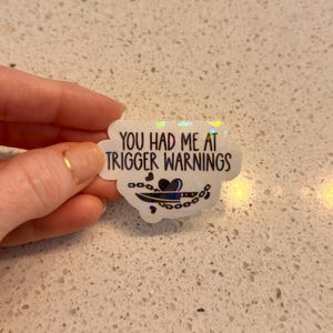 May include: A white sticker with the text "YOU HAD ME AT TRIGGER WARNINGS" in black. Below the text is a graphic of a knife, heart, and chain. The sticker has a holographic, iridescent finish.