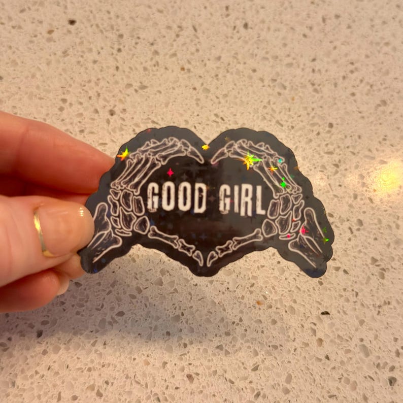 May include: A black sticker in the shape of a heart formed by skeleton hands. The words "GOOD GIRL" are in white block letters in the center. The sticker has a holographic, glittery effect.