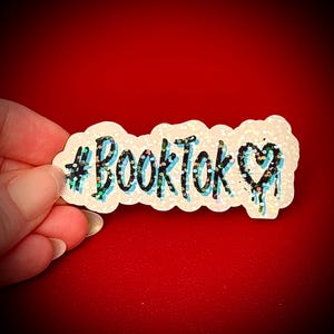 May include: A glittery sticker with the text "#BookTok" in black, outlined in light blue, and a dripping heart. The sticker is white with iridescent glitter and is held against a red background.