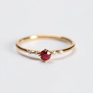 Puede incluir: Un delicado anillo de oro con una vibrante gema roja en el centro, flanqueada por piedras blancas más pequeñas y brillantes. La delgada banda del anillo y el contraste del oro y el rojo crean un diseño elegante. Una fina joya.