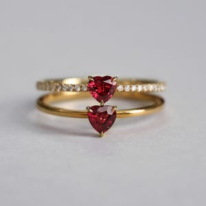 Puede incluir: Dos anillos de oro, uno con una banda de pequeños diamantes y el otro con una banda lisa. Cada anillo presenta una piedra preciosa roja oscura en forma de corazón. Los anillos están sobre un fondo suave y neutro.