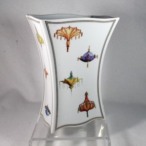 May include: White porcelain vase with a unique shape, featuring colorful, stylized parasol designs. The vase has a black and gold border and is displayed on a clear stand. The parasols are in shades of yellow, purple, green, and red.