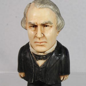 May include: A small, vintage figurine of a man with a stern expression. The figure has gray hair, a black suit, and a white shirt with a bow tie. The figurine is made of a light-colored material.