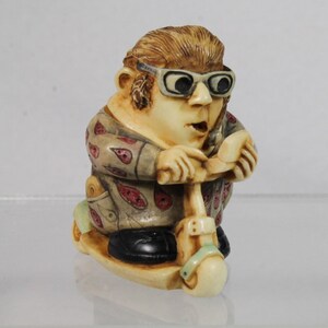 May include: A figurine of a person riding a scooter. The figure has light skin, brown hair, and is wearing glasses and a paisley patterned shirt. The scooter is cream-colored with a green accent.