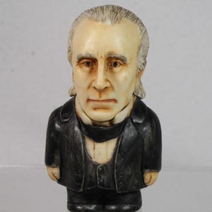 May include: A small figurine of a man with a serious expression, wearing a black suit and a white cravat. The figure has light skin, gray hair, and is standing upright. The figurine is a collectible item.
