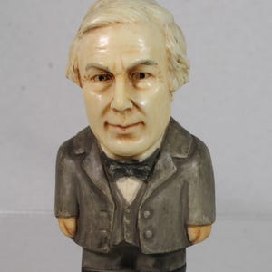 May include: A small, vintage figurine of a man in a gray suit with a black bow tie. The figurine has a cream-colored face and hair, and the suit has a slightly worn appearance. The figure is standing upright.